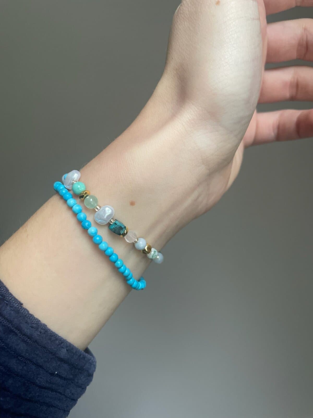 Zwei Armbänder am Handgelenk: eines aus türkisfarbenen Perlen, eines aus Perlen, Steinperlen und goldenen Details