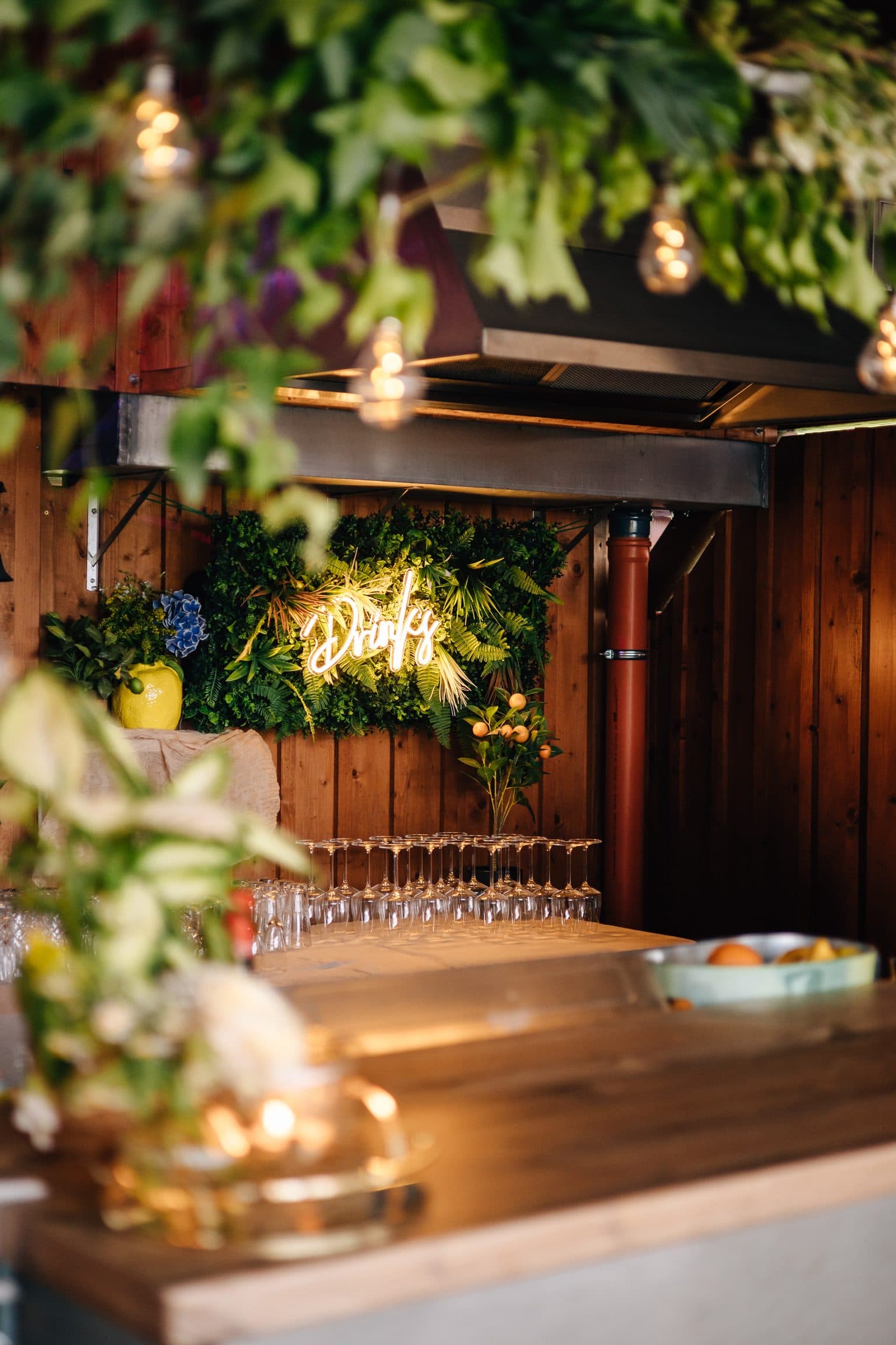 Getränkebar mit Neonschriftzug „Drinks“ vor einer grünen Pflanzenwand, Efeu und Glühbirnen-Lichterkette im Vordergrund