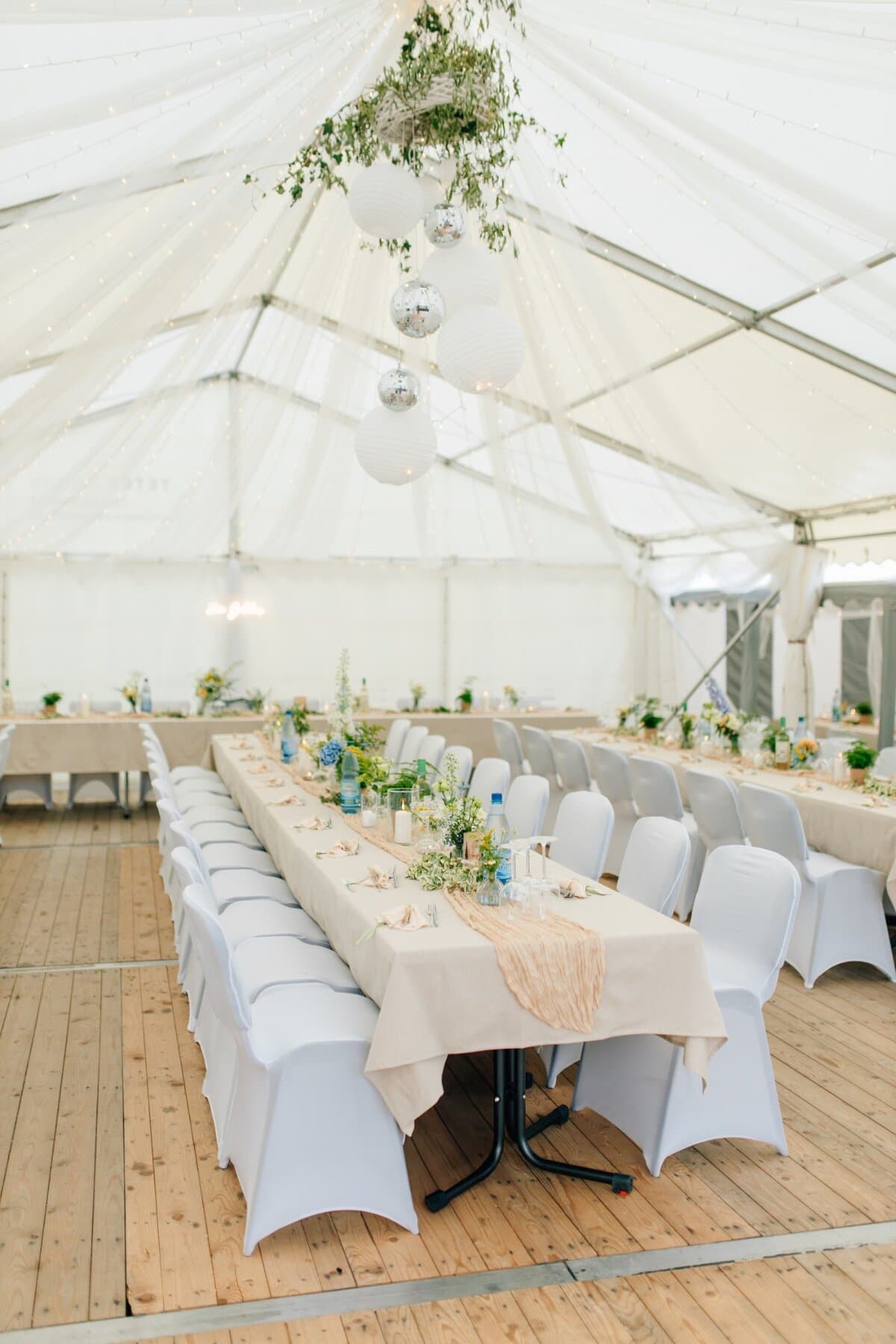 Festzelt mit weißen Stoffbahnen, Lichterketten, Papierlaternen und Diskokugeln über einer langen, floral gedeckten Tafel
