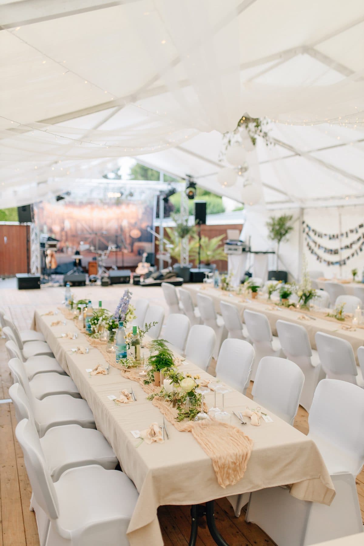 Festzelt mit langer, floral geschmückter Tafel, weißen Stühlen, Tüllbahnen unter der Decke und Bühne im Hintergrund