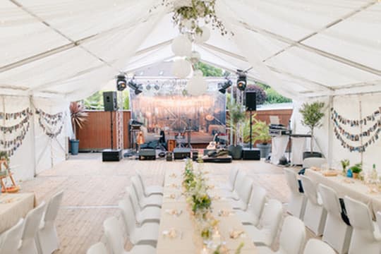 Festzelt mit langer, floral geschmückter Tafel, weißen Stühlen und Bühne im Hintergrund