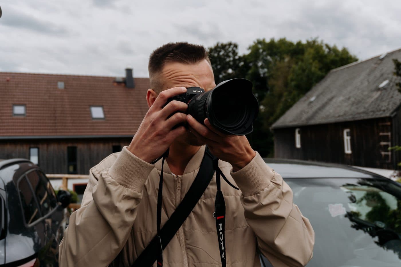 Fotograf mit Sony-Kamera in beiger Jacke vor einem Holzhaus