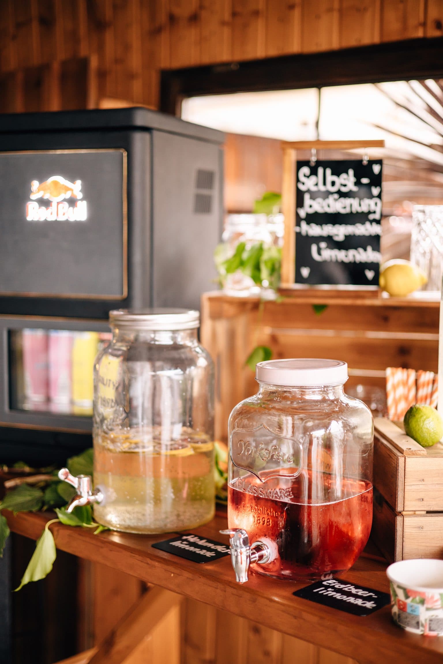Selbstbedienungs-Station mit hausgemachter Holunder- und Erdbeerlimonade in Glasspendern, Kreidetafel und Holzkisten