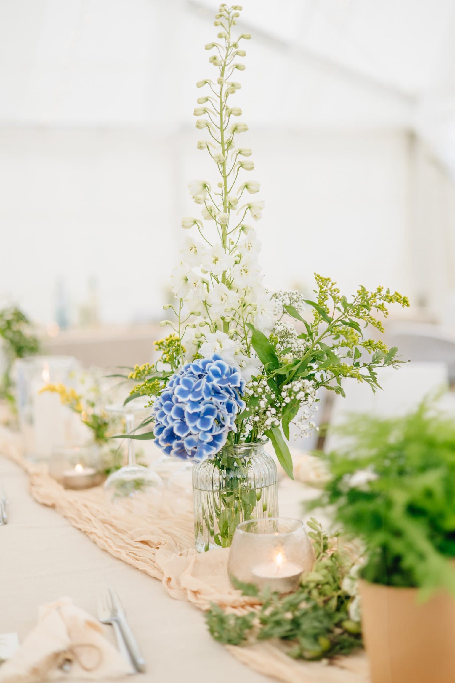 Tischgesteck mit blauer Hortensie, weißem Rittersporn und Schleierkraut in geriffelter Glasvase neben Teelichtern