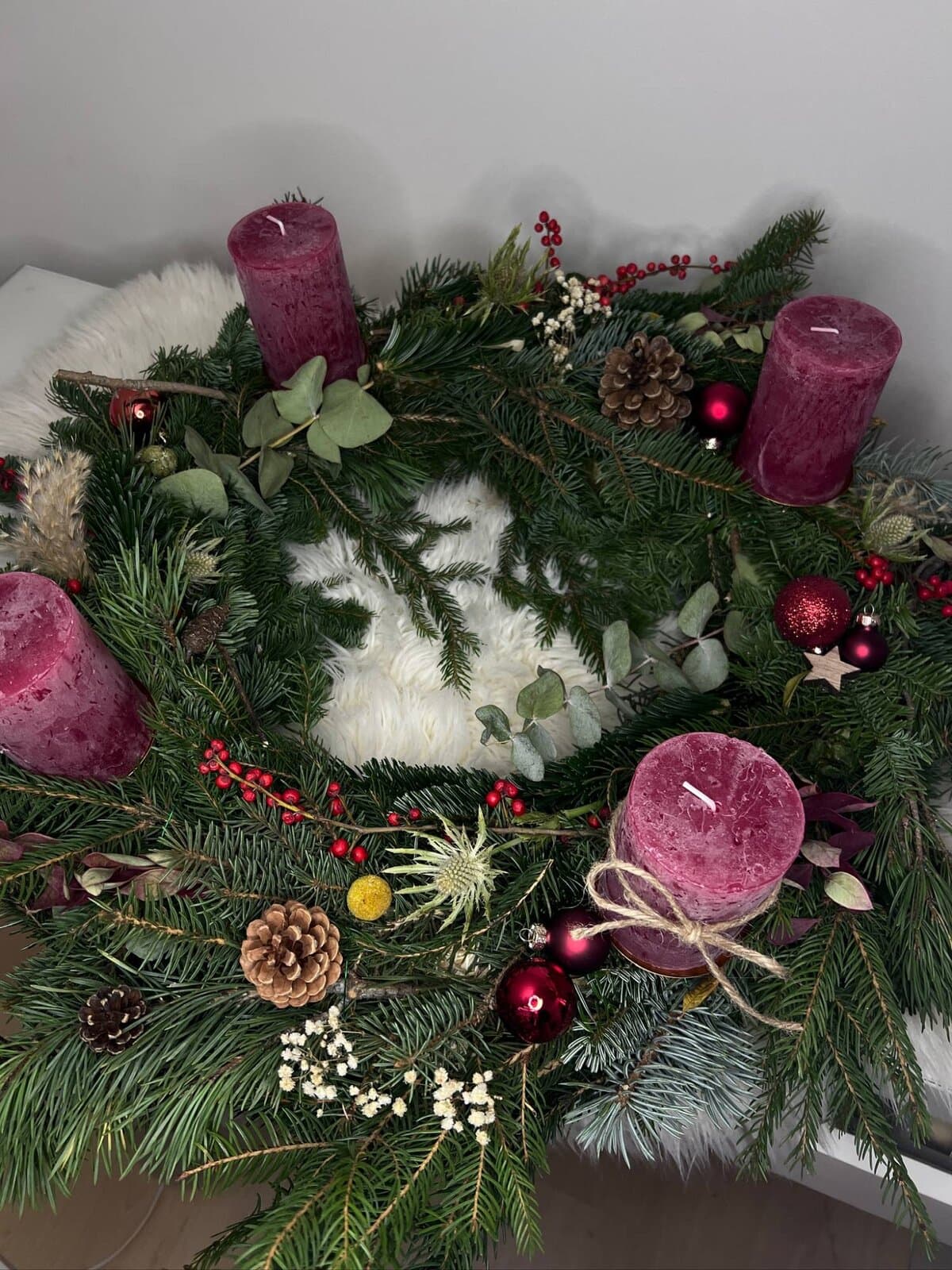 Adventskranz mit beerenroten Stumpenkerzen, Ilexbeeren, Disteln, Pampasgras und roten Kugeln auf einem weißen Fell