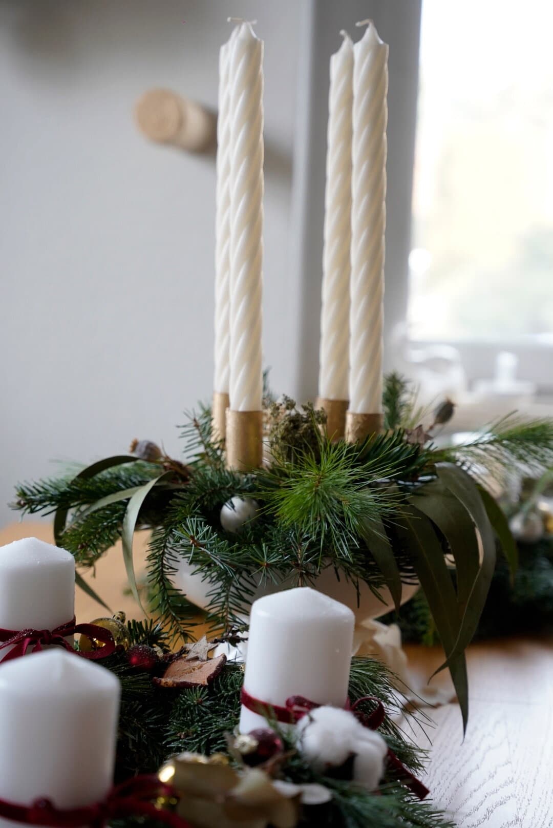 Adventskranz mit vier weißen gedrehten Stabkerzen in goldenen Haltern, Tanne, Eukalyptus und weißen Kerzen mit roter Samtschleife