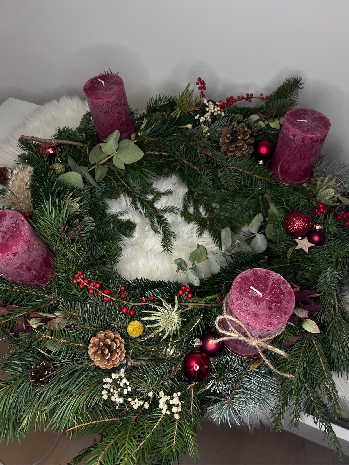 Handgebundener Adventskranz mit bordeauxroten Stumpenkerzen, Zapfen, Kugeln, Eukalyptus und roten Beeren auf einem weißen Fell