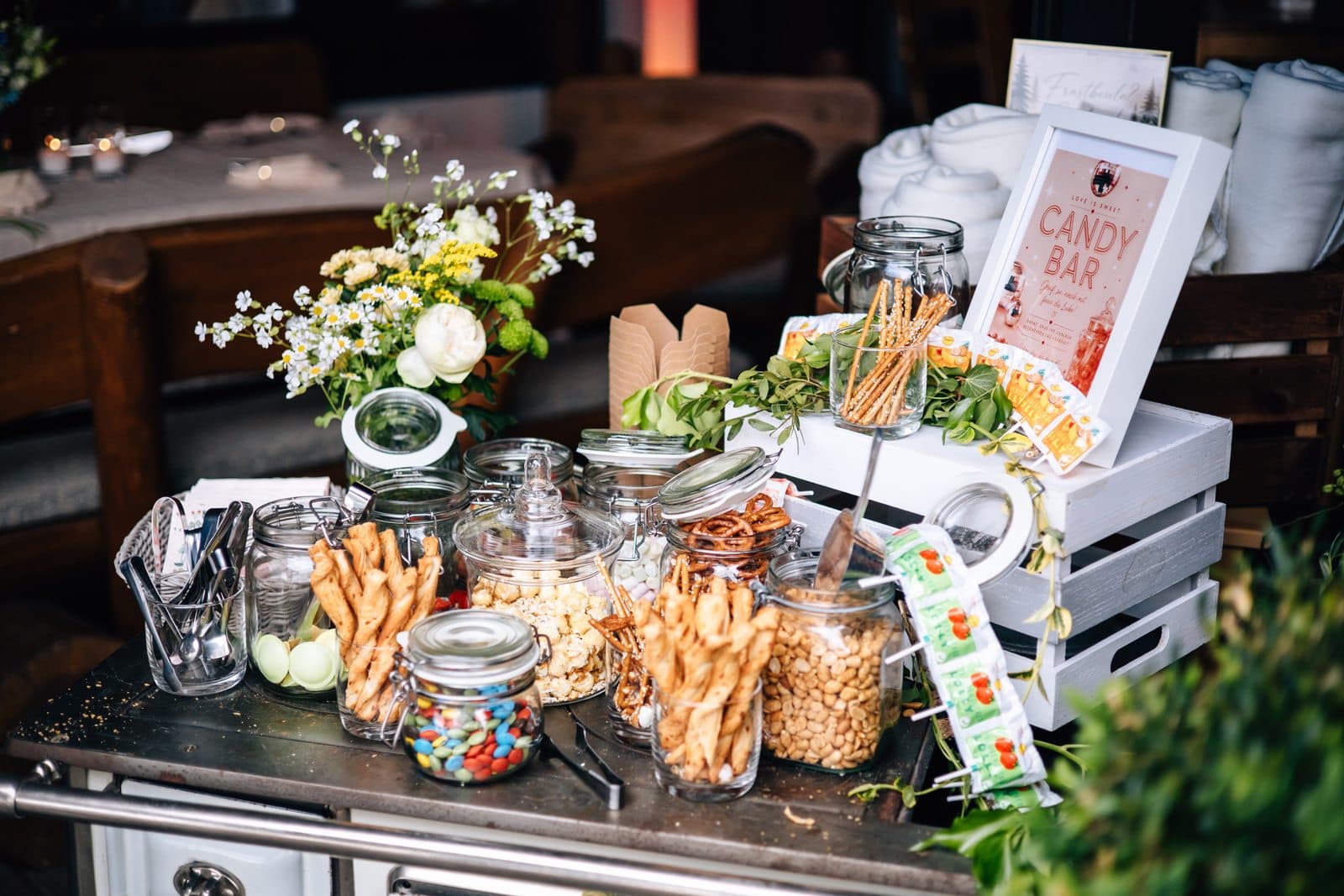 Candy Bar auf einem alten Herd mit Gläsern voller Popcorn, Erdnüssen, Salzstangen und Schokolinsen, dazu Wiesenblumen und ein Candy-Bar-Schild