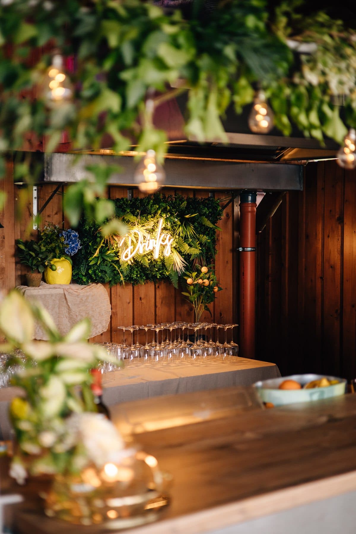 Drinks-Bar mit Neonschriftzug vor einer Pflanzenwand, Zitronenvase, Gläsern und Glühbirnen-Lichtern