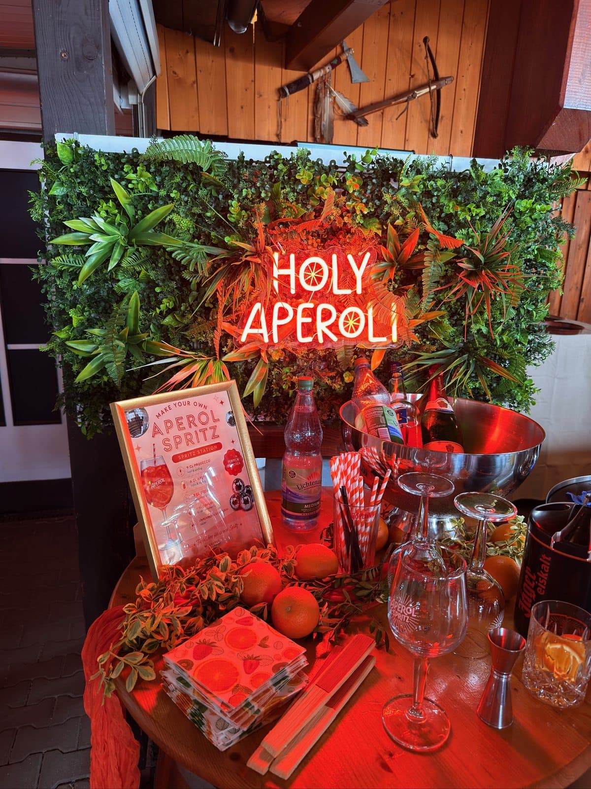 Aperol-Spritz-Station mit orangem Neonschriftzug vor einer Pflanzenwand, Orangen, Gläsern und Sektkühler