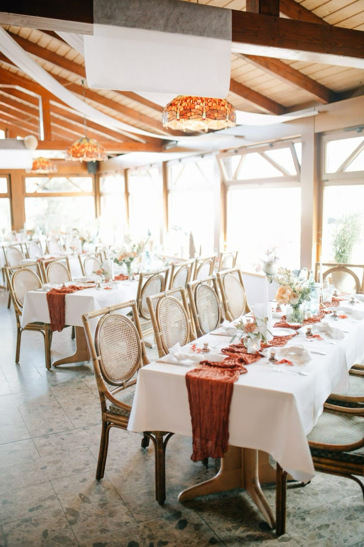 Hell gedeckte Hochzeitstafeln mit terrakottafarbenen Tischläufern und Rattanstühlen unter Holzbalken