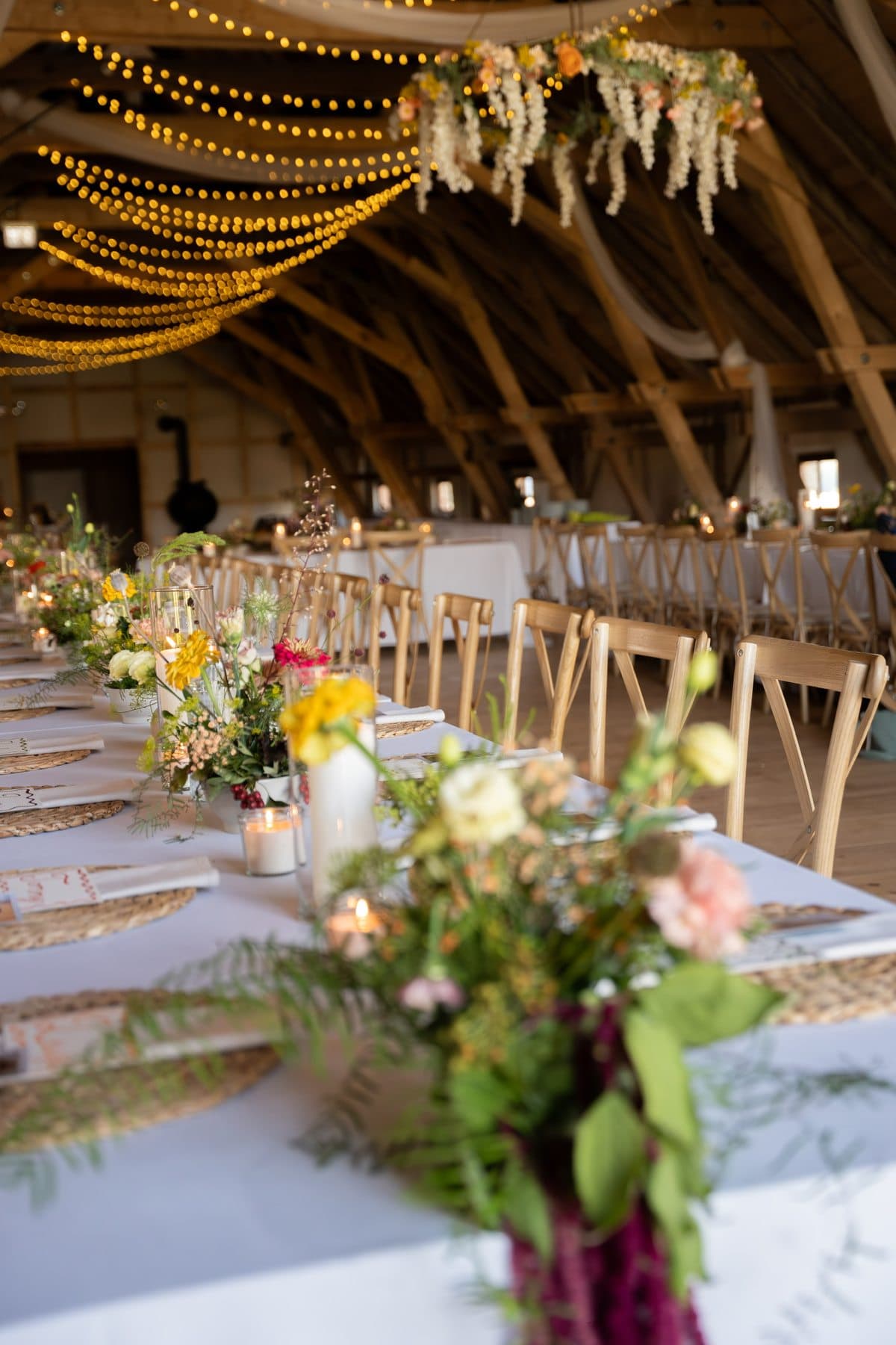 Lange Hochzeitstafel in einer Holzscheune mit Lichterketten, Blumenstrauß und hängendem Blumenschmuck